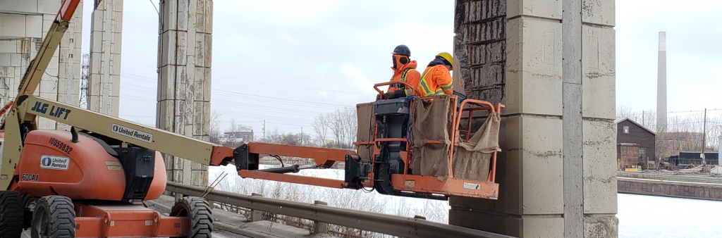 Construction workers in a cherry picker lift performing maintenance work on a Gardiner bent