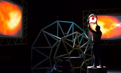 Photo credit: Michael Kast. Person on sage. Holding circle. near dome structure. Fire projections on screens.