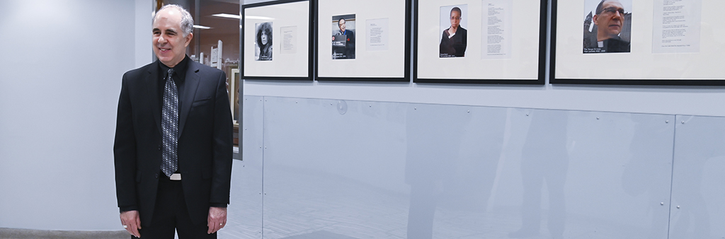 Al Moritz stands in hallway with wall lined with portraits and poems from all previous Toronto Poets Laureate