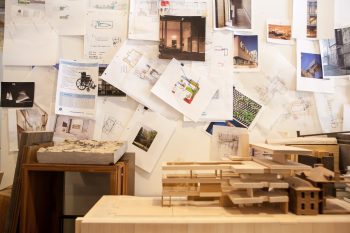 Studio with a wall filled with architectural drawings and photos. In front of this, small models of a building are placed on a table.