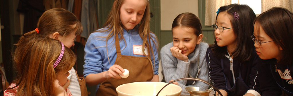 Photo of children standing around a table in aprons cracking eggs and making batter.