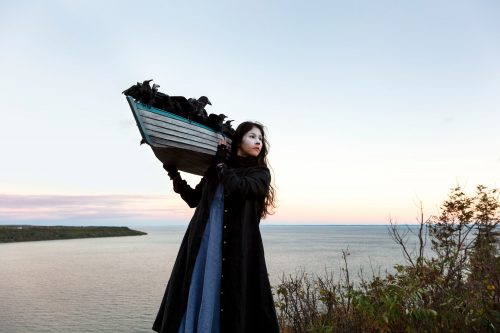 Woman with small boat on her shoulder. water,m vast sky in background.
