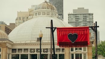 A banner from the Red Embers installation hanging in front of the Allan Gardens Conservatory. The flag is red and has a black heart on it. It is hung on charred-black gates.