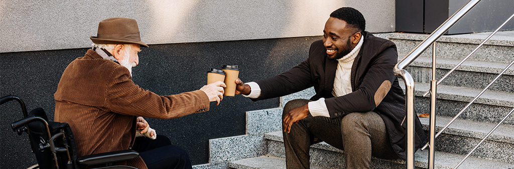 Man sitting on steps and elderly man sitting in a wheelchair cheers coffee cups with a smile.