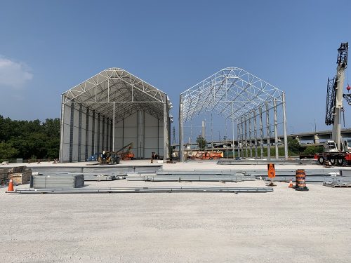 Image of Gardiner Expressway Strategic Rehabilitation, Jarvis to Cherry fabrication site located on lands near Lakeshore Blvd E and Cherry St. Image features two large tent structures in the process of assembly straight on view, one with a canvas cover and the other just a metal frame with the Gardiner Expressway in the background.