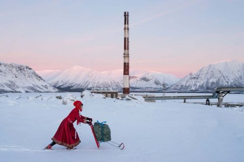 Image info: Marja Helander, Dolastallat (To have a campfire), 2016. Video still. Courtesy of the artist.. woman in red coat pushing sleigh. snow on ground. mountains, smoke stack in background.