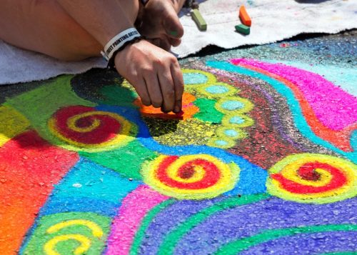 Close-up of person's hand drawing a colourful mural on pavement.