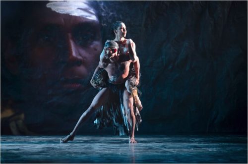 Image info: Bangarra Dance Theatre. Photo: Daniel Boud and Jacob Nash. two performers standing, one on back of other. Large face in background.