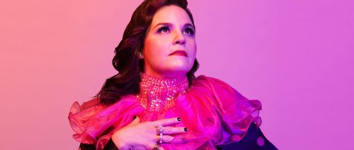 Image info: photo of Michaela Washburn by Tanja-Tiziana. Pink background, performer with pink and sequined neck ruffle looking off into distance