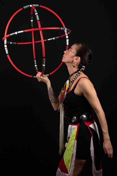 Image info: Barbara Diabo, by Mario Faubert. Performer in costume holding red hoops aloft.