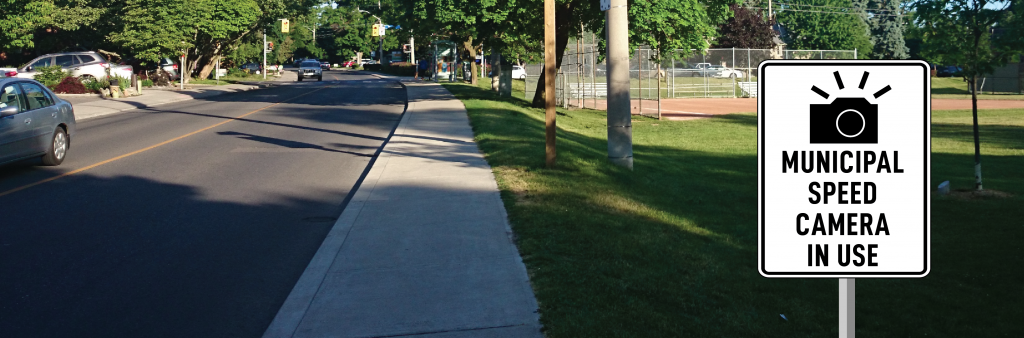 A image of a street with a school to the right side, with a sign reading municipal speed camera in use.