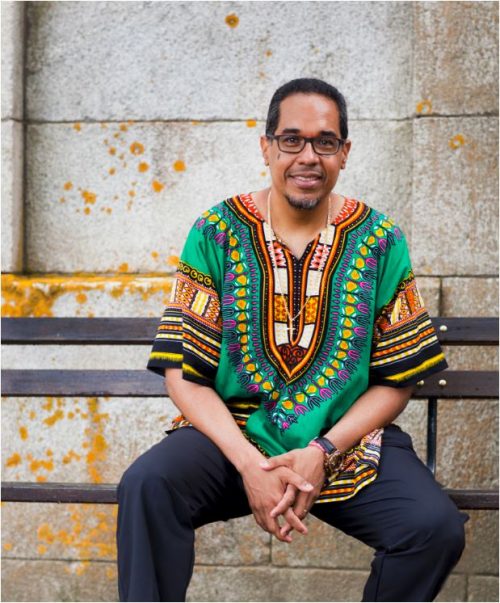 Image info: Danilo Pérez. By John Abbott. Performer sitting on bench in front of stone wall wearing green, patterned shirt