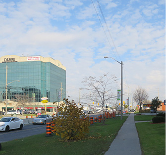 Photo of Finch Ave facing East with traffic heading west, with sidewalks and a mid-rise office building with retail on the ground floor.