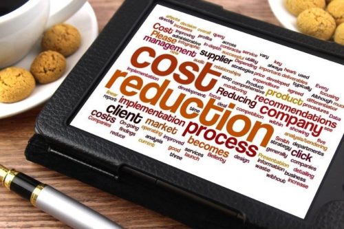Tablet resting on a table next to a coffee cup and plates of cookies with a word cloud displayed onscreen highlighting the words cost reduction, process and implementation.