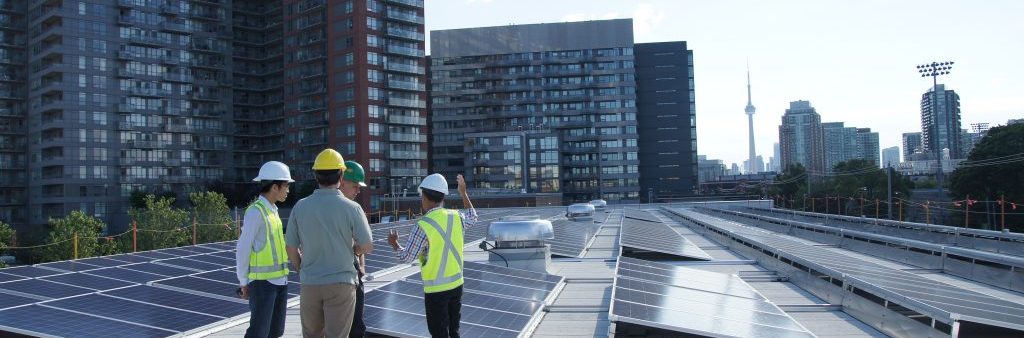 An image of four City of Toronto workers in hardhats on a building roof with rows solar PV panels. The CN Tower can be screen on the horizon