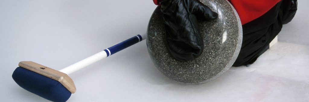 Someone holds a curling rock. A broom sits on the ice.