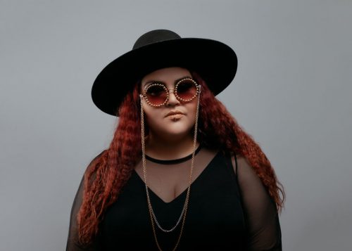 Portrait of DJ KOOKUM in sun glasses and a black hat against grey background.