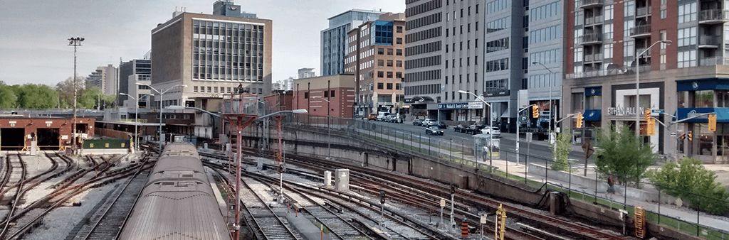 A photo of a northbound subway train proceeding through the Davisville Yard, with mixed-use buildings along Yonge Street visible in the background.