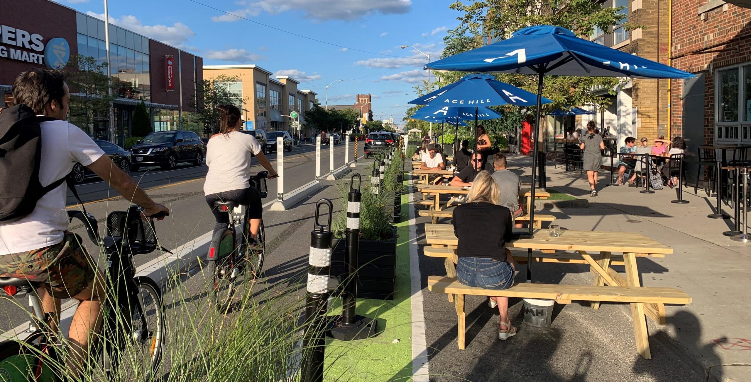 Image of Danforth Avenue after the bike lane installation with painted bike lanes separated by planters, bollards and concrete barriers. To the right of the bike lanes are curb lane patios with seating.