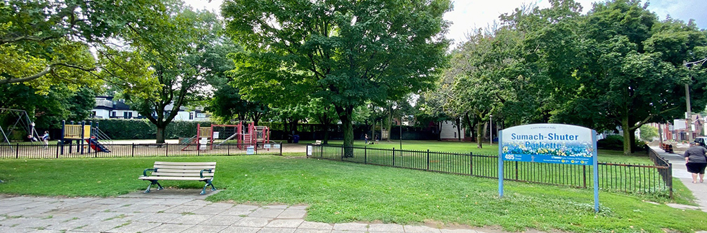 Photograph of Sumach-Shuter Parkette. In the background are large trees and colouful playground equipment. In the foreground is a bench and a path on a grassy area. On the right is a City of Toronto park sign that reads Sumach-Shuter Parkette.