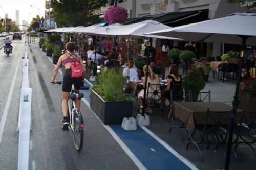 Person riding bicycle in a protected bike lane passing by a cafe patio. The cafe and bike lane are seperated by a painted buffer and planter box.