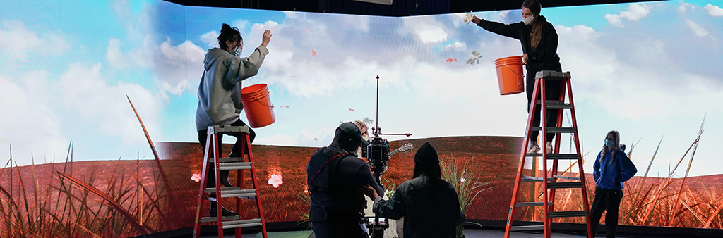 Image of film shoot taken from behind the camera, with technicians at top of ladders dropping brown leaves on actor.