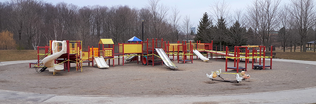 A picture of Milliken Park Playground on a day with overcast weather. The playground has a circular footprint with sand under all play equipment and a narrow concrete path surrounding the playground. The playground is larges, with many different climbing features and play panels and is surrounded by mature trees and open green space.