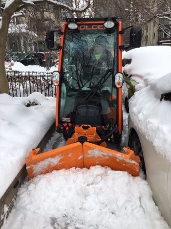 A small plow clearing snow from a tight space between a retaining wall and a parked car.