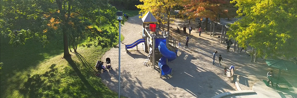 Aerial view of the playground in Willowdale Park. The playground features a large play structure with multiple slides, including a blue spiral slide a straight blue slide. The play structure has a small, roofed area and is surrounding by trees with green, yellow, and red foliage indicating it’s fall season. There are several people using the playground, including children playing and adults supervising.