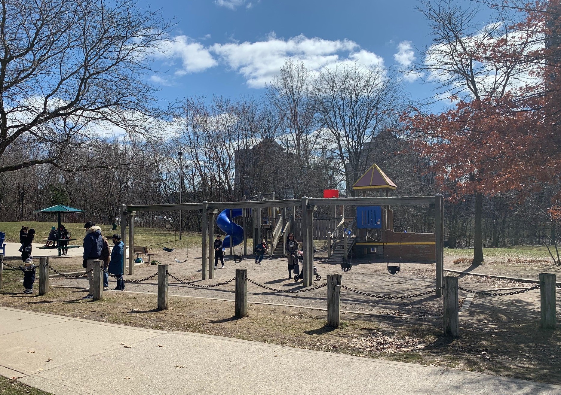 A photograph of the playground taken on a sunny winter day. The playground has post chain fencing on one side parallel to the three swing sets on top of sand play surfacing. 