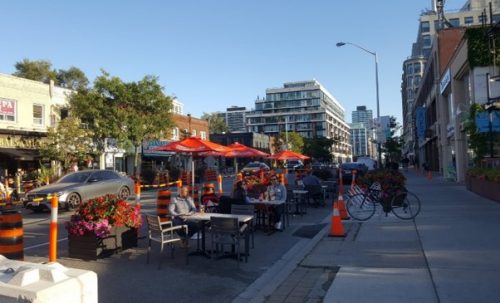 The curb lane shows a row of patio cafes being enjoyed by people on a sunny day.