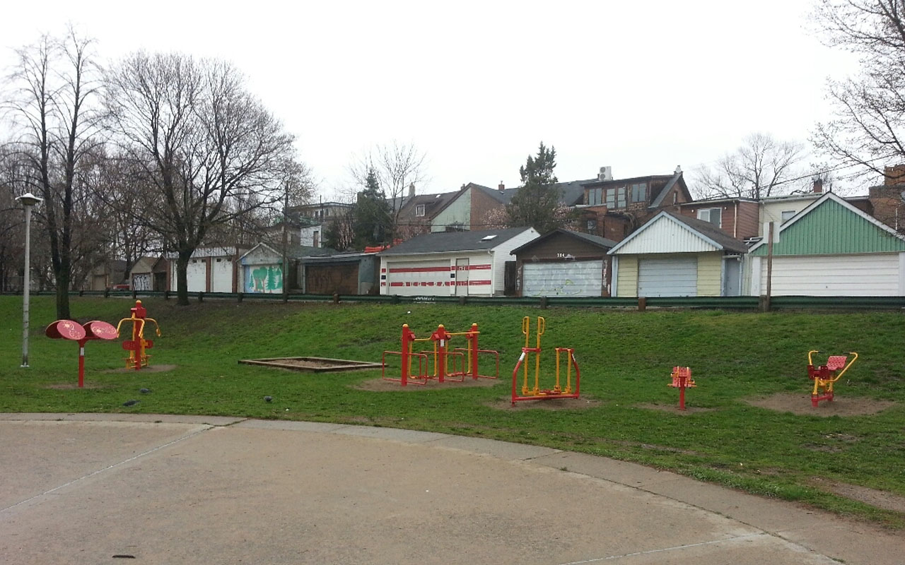 An image of some of the stand-alone play elements surrounding the wading pool. The play elements sit on top of grass, are red and yellow in colour and include a small sand box, play panels and see-saw. 