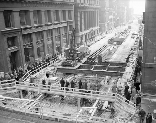 Workers place wooden planks over an excavation to make a roadway. Crowds on both sides of the street are watching them.