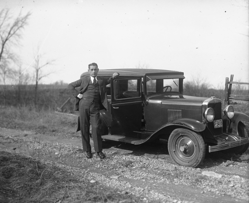 A man is standing in front of an automobile on a dirt road. His hand is resting on the open door.