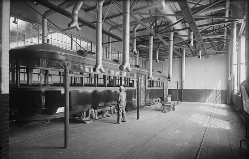 The room is narrow, and sunlight is coming in through the wide windows. A worker is spraying glossy paint onto the side of a streetcar.