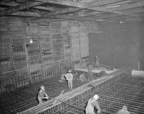 Workers are installing metal rebar along the floor and walls of a large underground room.
