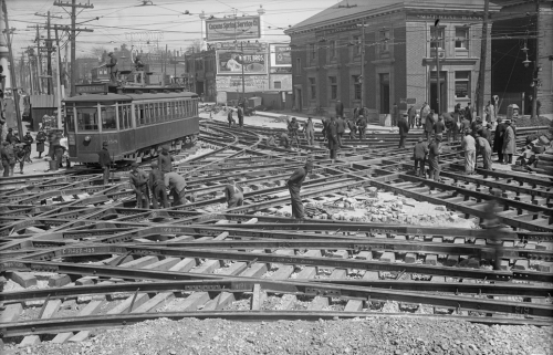 Many men digging the road up around a network of streetcar tracks going in every direction.