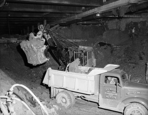 In an underground space, the bucket of an excavating machine moves dug-up earth into the back of a dump truck.