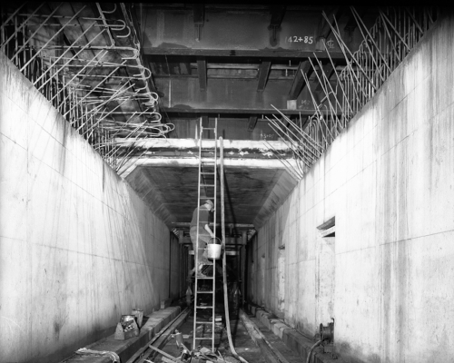 A worker in a square concrete tunnel carries a bucket up a ladder to a second level.