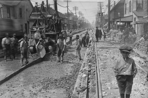 A boy in knee-length pants and a cap stands with his hands in his pockets. Behind him, men are constructing a road.