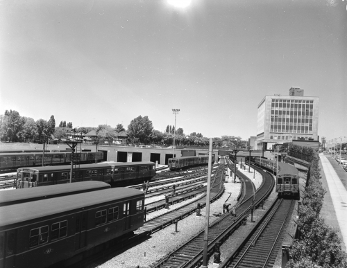 Several subway trains sit on tracks. In the distance is a square, modernist office building with many windows.