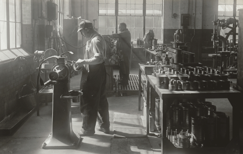 A worker is polishing equipment parts on a lathe. Behind him is a table filled with cylindrical metal parts.