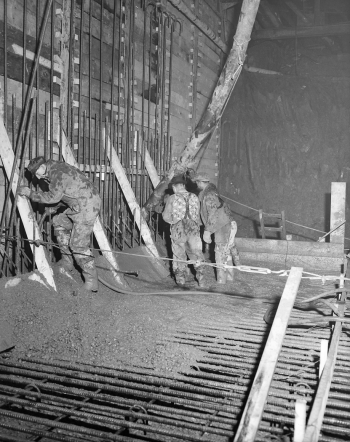 Workers are holding a large pipe as it pours concrete onto a wall and floor covered in a mesh of metal rebar.