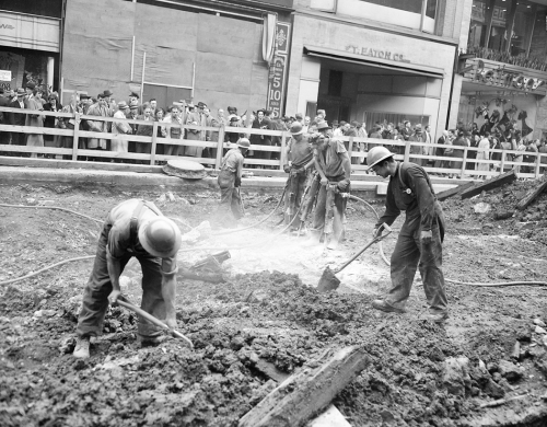 Workers dig in the ground with shovels and jackhammers. In the background, people are lined up behind a wooden fence, watching.
