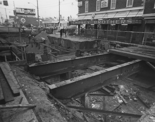 Beams over excavated area. Workers inside fenced area near hole. Dempsey Brothers store and gas station in background.