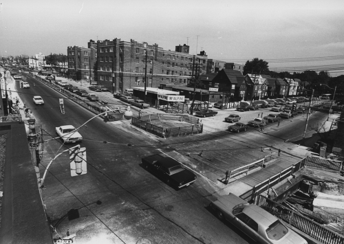 McAlphine sign at construction site. Traffic in streets. Background shows apartment building and houses along street.