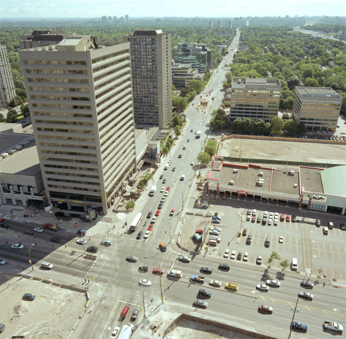 Traffic on roads, office towers, and shopping centre with parking lot. Background shows green space.