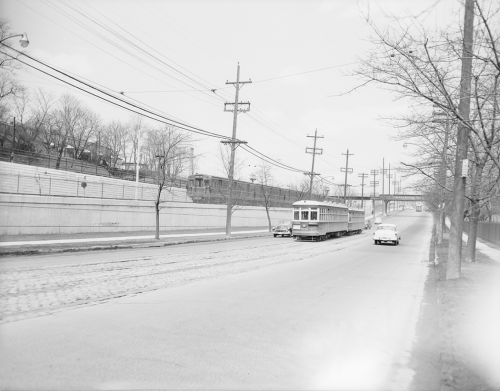 An older streetcar drives up a street, while a subway train can be seen on an embankment behind it.