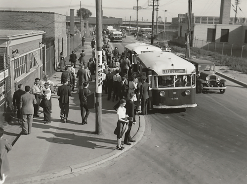 Crowds of people awaiting buses in an industrial area.