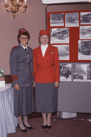 Two uniformed women standing near display. One woman is wearing a grey uniform with burgundy details. The other woman is wearing a grey skirt, white blouse and red blazer with a red cap.